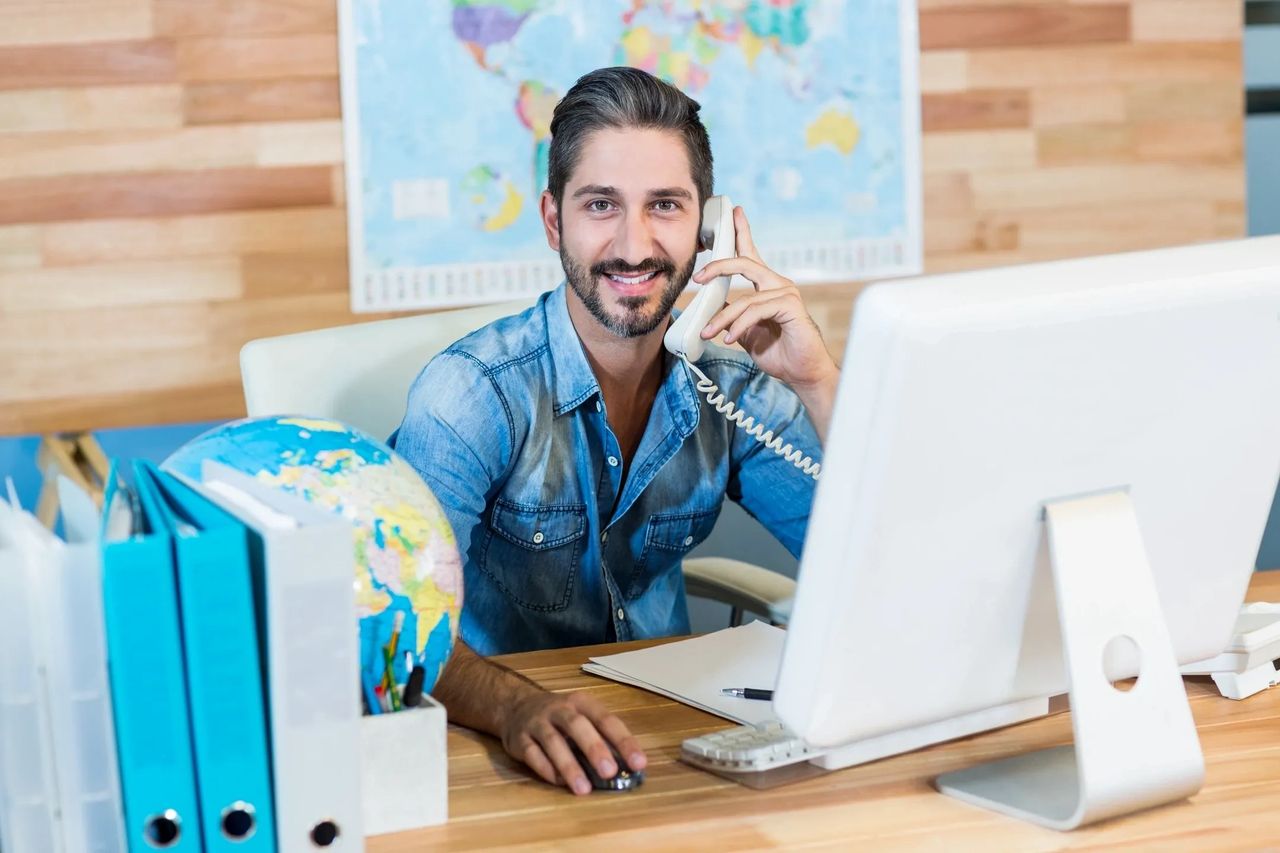 Bearded man at desk on a phone call, with map of the world behind him